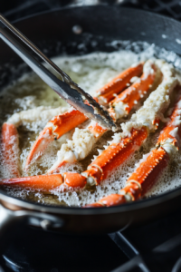 A top-down view of coated crab legs being carefully placed into a large skillet or deep fryer with hot oil on a stovetop. The crab legs are beginning to turn golden brown as they fry, with warm natural light capturing the sizzle and crisping process. The scene is clean and focused, emphasizing the frying process.