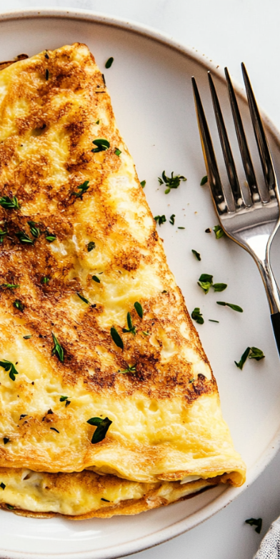A top-down view of a golden, fluffy cheese omelette served on a white ceramic plate, placed on a white marble countertop. The omelette is folded in half, revealing melted cheese inside, and garnished with finely chopped fresh herbs. The plate is accompanied by a fork and knife, neatly placed beside it. The background is simple and clean, ensuring the focus remains on the rich texture and appealing look of the omelette. This dish is perfect for a quick breakfast or brunch, offering a soft, creamy interior with a slight crisp on the edges.