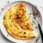 A top-down view of a golden, fluffy cheese omelette served on a white ceramic plate, placed on a white marble countertop. The omelette is folded in half, revealing melted cheese inside, and garnished with finely chopped fresh herbs. The plate is accompanied by a fork and knife, neatly placed beside it. The background is simple and clean, ensuring the focus remains on the rich texture and appealing look of the omelette. This dish is perfect for a quick breakfast or brunch, offering a soft, creamy interior with a slight crisp on the edges.