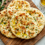 A top-down view of freshly baked cottage cheese flatbreads on a wooden board or platter on a marble countertop. The flatbreads are golden brown with a light, crispy texture, drizzled with olive oil or sprinkled with fresh herbs, and the scene is clean and inviting.
