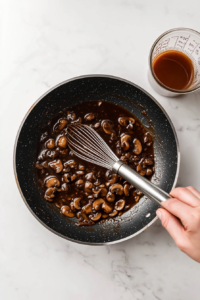 This image shows shallots and garlic being added to the sautéed mushrooms in a pan, with the mixture sizzling as the base for the marsala wine sauce is prepared.