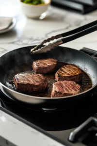 This image shows filet mignon steaks being seared in a large skillet with olive oil, creating a golden-brown crust on each side before finishing in the oven.