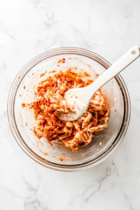 This image captures spicy mayonnaise-coated imitation crab sticks inside a clear glass bowl, tossed and mixed using a white bamboo rice paddle. The creamy orange sauce clings to the crab strips evenly. The bowl is placed on a clean white marble surface. The vibrant colors and textures highlight the preparation step essential for creating delicious sushi fillings.