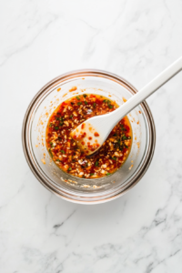 This top-down view shows spicy chili sauce and creamy mayonnaise being stirred together in a clear glass bowl using a white bamboo rice paddle. The mixture has a rich orange color and is being blended smoothly. The bowl is set on a clean white marble countertop, creating a bright and minimal background that enhances the visual appeal of the simple yet flavorful sauce preparation.