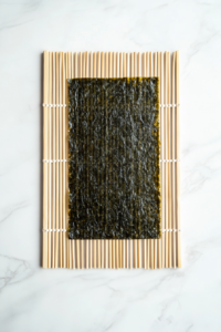 This photo features a top-down view of a single sheet of glossy nori seaweed placed on top of a traditional bamboo sushi rolling mat. The setup is positioned on a bright white marble countertop, with no distractions in the background. The focus is on the shiny nori, ready to be topped with sushi rice and other ingredients in the following steps.