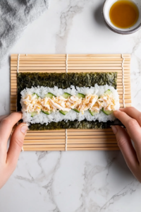 A clean top-down shot showing hands rolling sushi tightly with a bamboo mat placed over a white marble surface. The edges of the nori show fillings of sushi rice, spicy crab meat, and cucumber. The rolling process is captured mid-action, emphasizing the technique and alignment necessary for making a perfectly formed sushi roll. The setup remains minimal and focused on the rolling process.