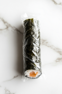 This image captures a completed sushi roll tightly wrapped in clear plastic wrap, resting on a bright white marble countertop. The roll appears compact and uniform, with the dark green nori forming a seamless outer layer. The setup is clean and uncluttered, emphasizing the chilling step that helps firm up the roll before slicing and serving the final sushi pieces.