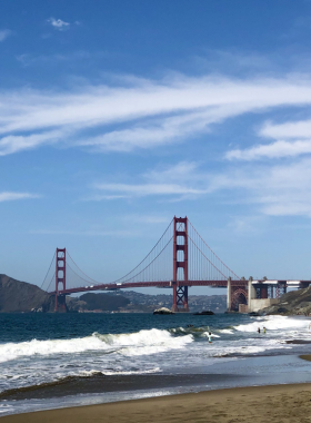Baker_Beach_Golden_Gate.jpg A stunning view of Baker Beach with the Golden Gate Bridge in the background. The sandy beach is dotted with sunbathers and beachgoers enjoying the warm sun, while gentle waves lap at the shore under a clear blue sky.