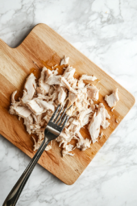 Cooked chicken breasts being shredded with a fork on a wooden cutting board.