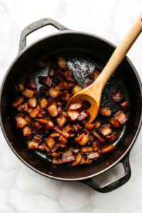 Diced thick-cut bacon sizzling in a large pot, turning golden and crispy.