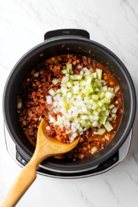 Diced onions and sliced celery cooking in a pot with rendered bacon fat, with minced garlic and seasonings being stirred in.