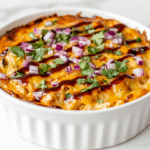 side view of a golden brown BBQ chicken dip served in a white ceramic baking dish. The bubbly cheese topping is perfectly melted and finished with a drizzle of BBQ sauce, chopped red onion, and fresh cilantro. The dish sits on a white marble surface and is ready to be served with chips or crackers.