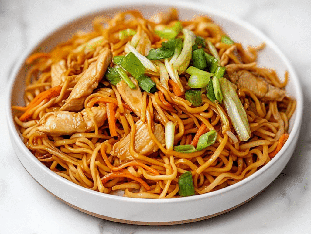 side view of the final chicken chow mein dish served on a white ceramic plate placed on a clean white marble countertop. The noodles are cooked perfectly and coated in a glossy sauce, surrounded by vibrant vegetables like cabbage, carrots, and green onions. Thin chicken strips are spread evenly, making the dish colorful and delicious. The composition is simple, highlighting the textures and colors of the completed chow mein.