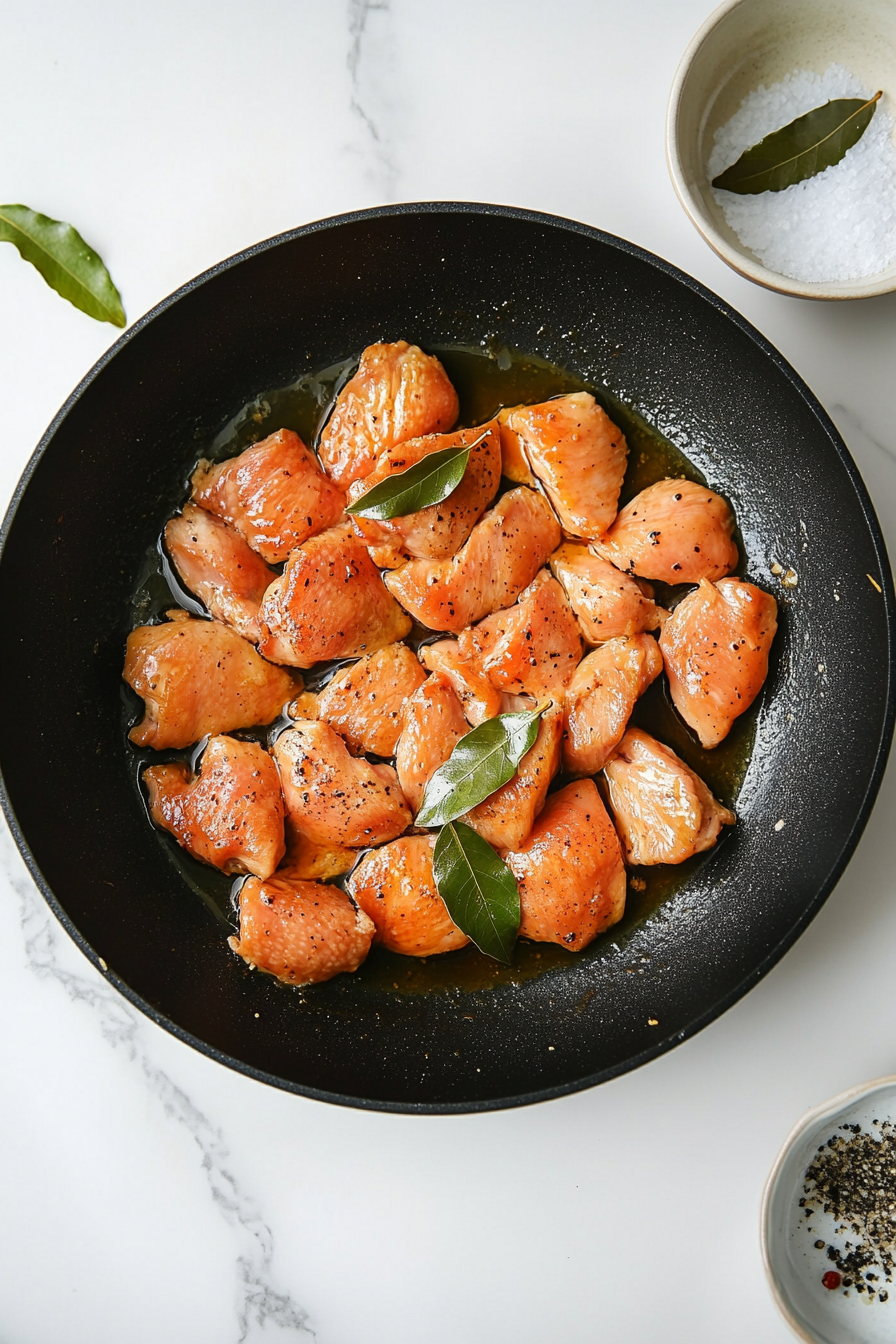 A top-down shot of marinated chicken thighs sizzling in a black skillet over a white marble countertop. The chicken is seared to a golden brown while bits of garlic and bay leaves remain visible in the pan. This image captures the start of flavor development through the browning process, offering a clear view of how the dish begins to build its rich taste. A reserved marinade bowl and wooden spatula add practical context.